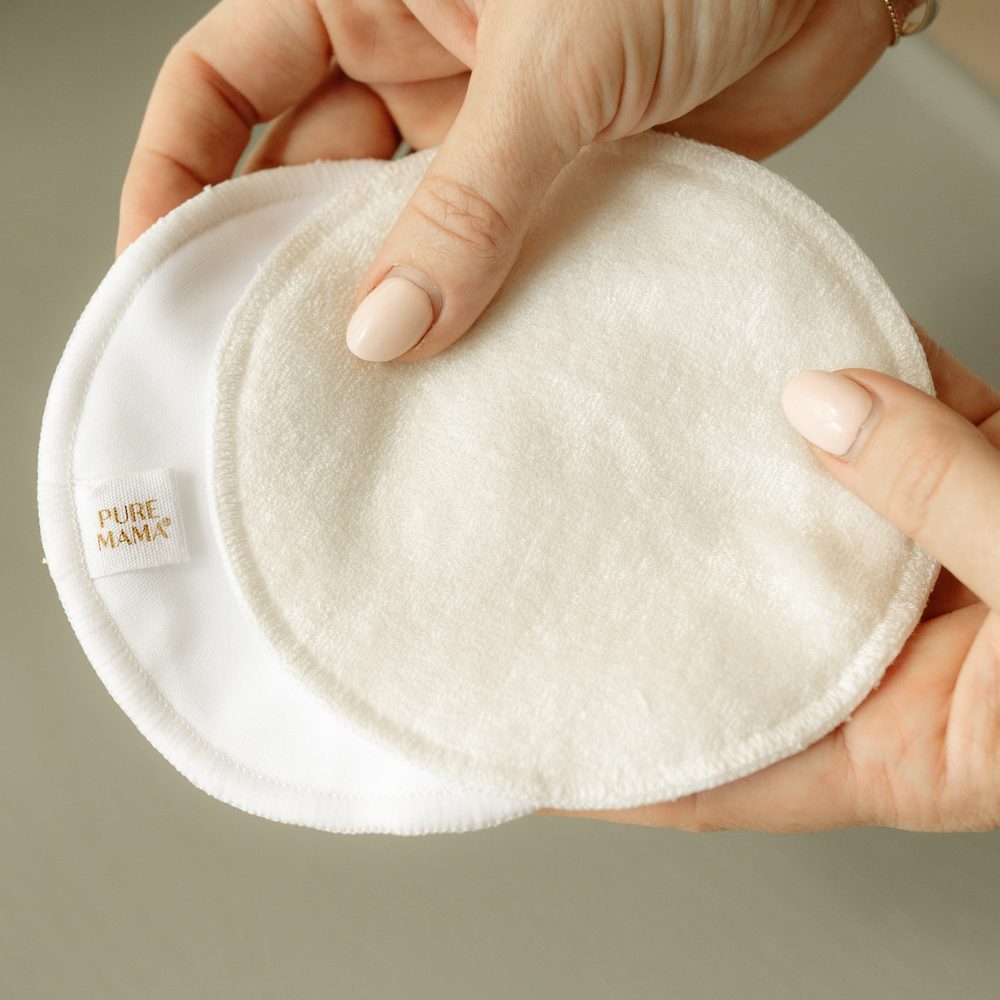 A person with manicured nails holds two round, soft PURE MAMA Skincare Nursing Pads (set), one displaying a small “PURE MAMA” tag, against a neutral, out-of-focus background.