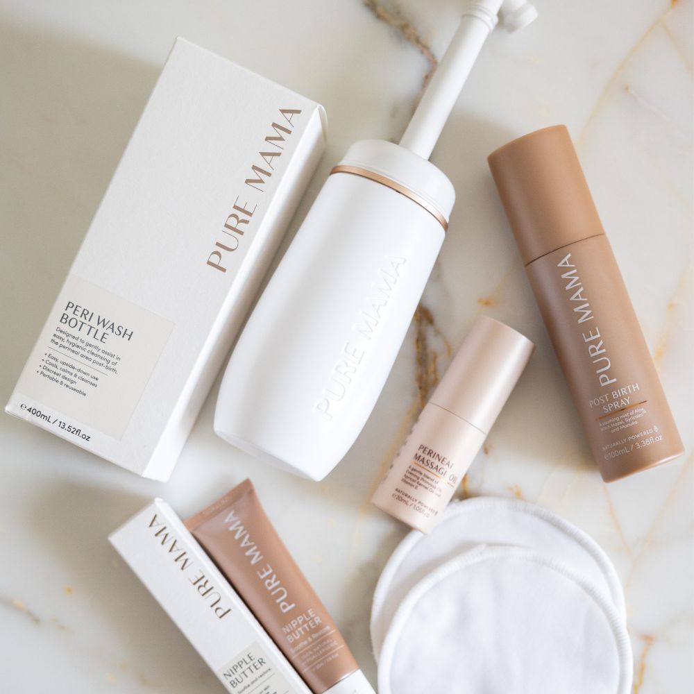 Flat lay of the Pure Mama NZ Birth & Postpartum Recovery Kit, featuring a peri wash bottle, perineal massage oil, tubes, a small bottle, product box, and reusable cotton pads on a white marble surface.