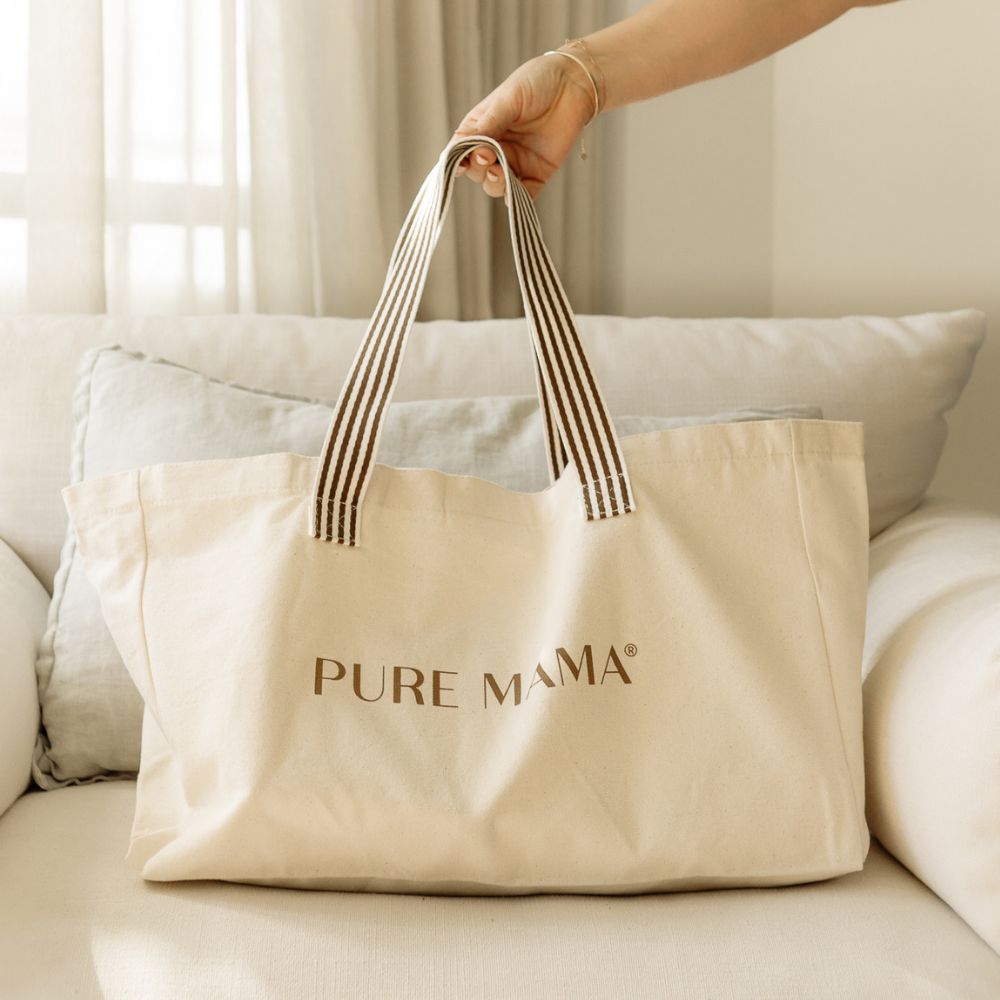 A large cream-colored cotton tote bag featuring PURE MAMA Skincare in brown letters sits stylishly on a light-colored sofa. Striped handles enhance its design, while natural light streaming through curtains adds a serene backdrop.
