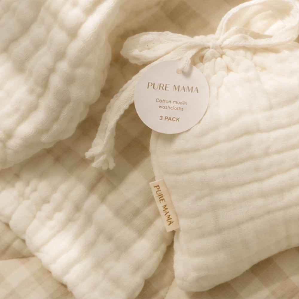 Three folded cream-colored cotton muslin washcloths by Pure Mama, tied with a drawstring and accompanied by a round paper tag, arranged on a beige and white gingham fabric background.