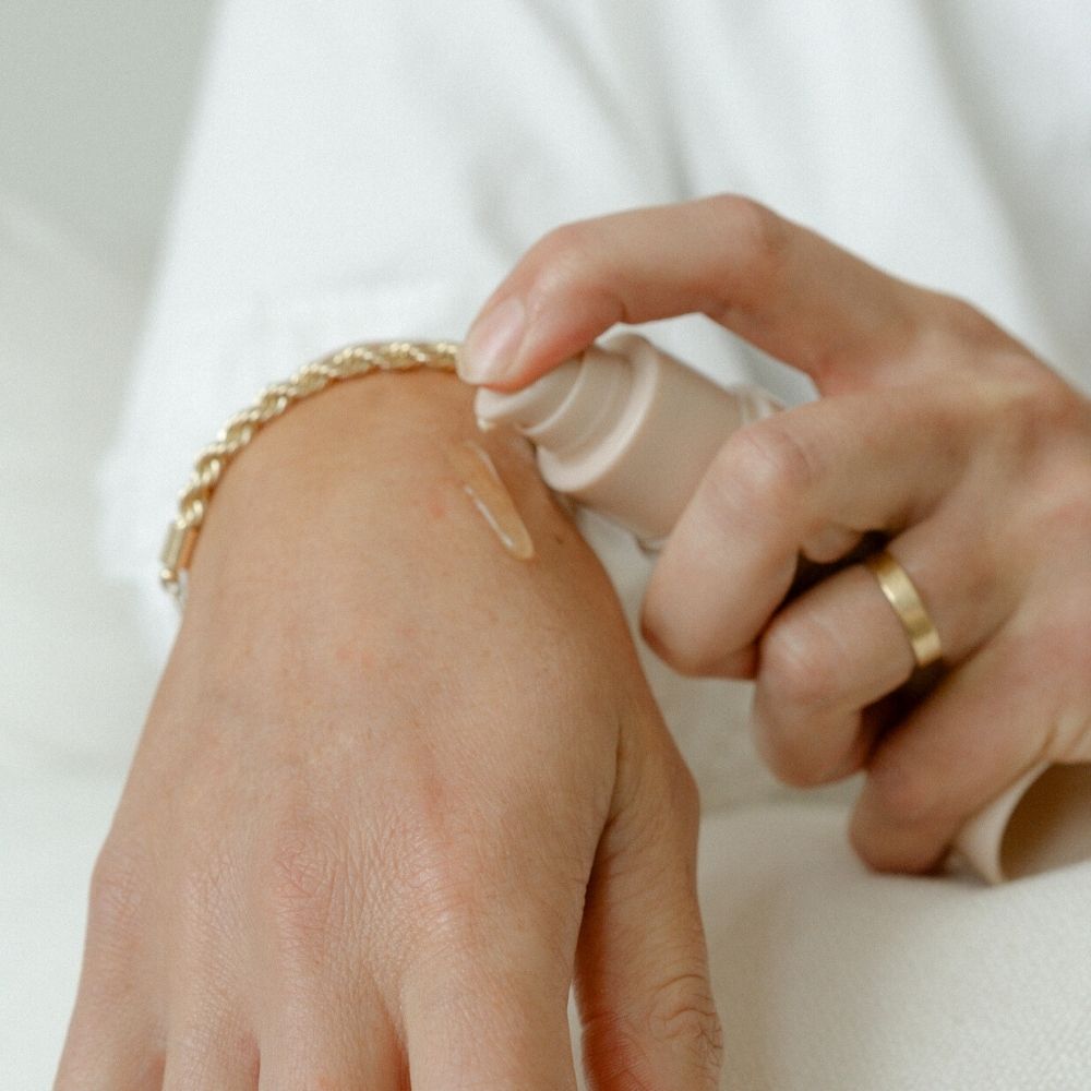 A person dispenses Pure Mama NZ Perineal Massage Oil onto the back of their hand to show skin elasticity. They wear a gold bracelet and ring, with a light, neutral background.