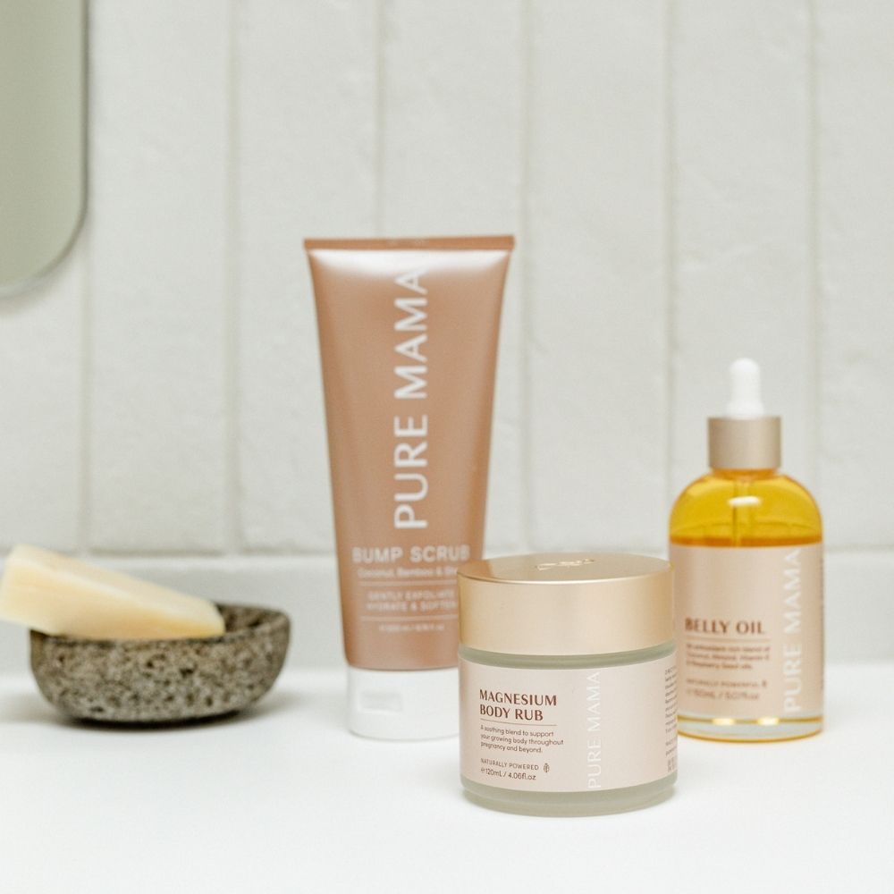 The Pure Mama NZ Pregnancy Care Set, featuring Bump Scrub, Belly Oil, and Magnesium Body Rub, is displayed on a bathroom countertop beside a soap bar in a small bowl, with off-white tiled walls in the background.