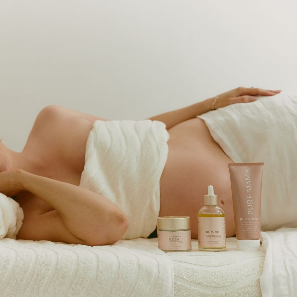 A pregnant person reclines on their side, draped in a white towel. In the foreground are three skincare products—a jar, a serum bottle with a dropper, and a tube—all part of Pure Mama NZs Pregnancy Care Set. The ambiance is serene with a gentle, neutral background.