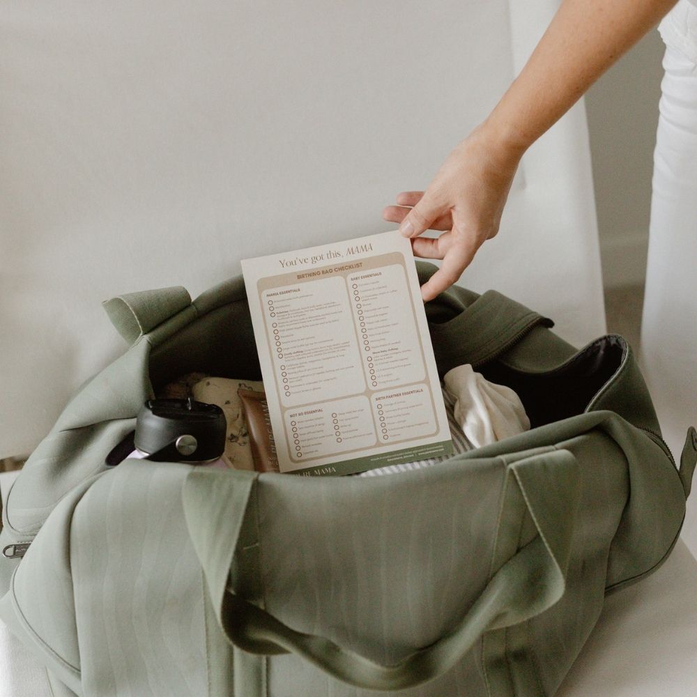 A person packs a Pure Mama NZ Birthing Bag Checklist into a green duffel with clothing and a black water bottle—making it easy to organize essentials quickly.