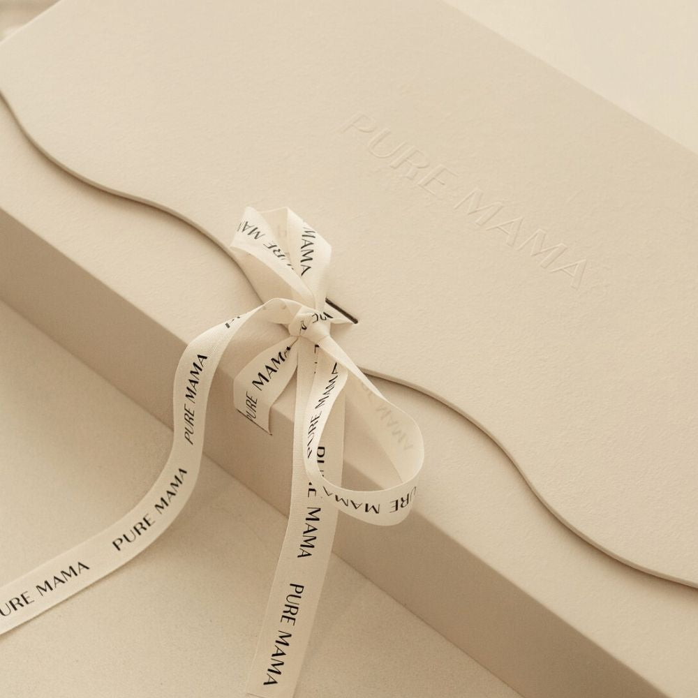 A cream gift box embossed with the Pure Mama NZ logo and wrapped in a matching ribbon inscribed PURE MAMA, paired with a Pure Mama Gift Voucher, displayed on a neutral background.