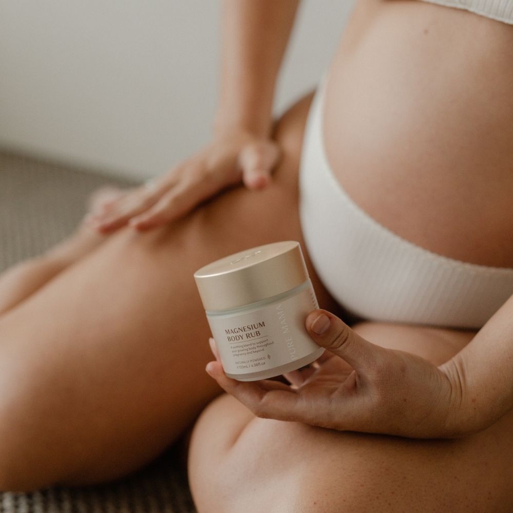 A person in white underwear sits on the floor, holding a jar of Pure Mama NZ Magnesium Body Rub from the Pregnancy Care Set, with their other hand on their thigh and their pregnant belly visible. The softly blurred background adds focus to the scene.