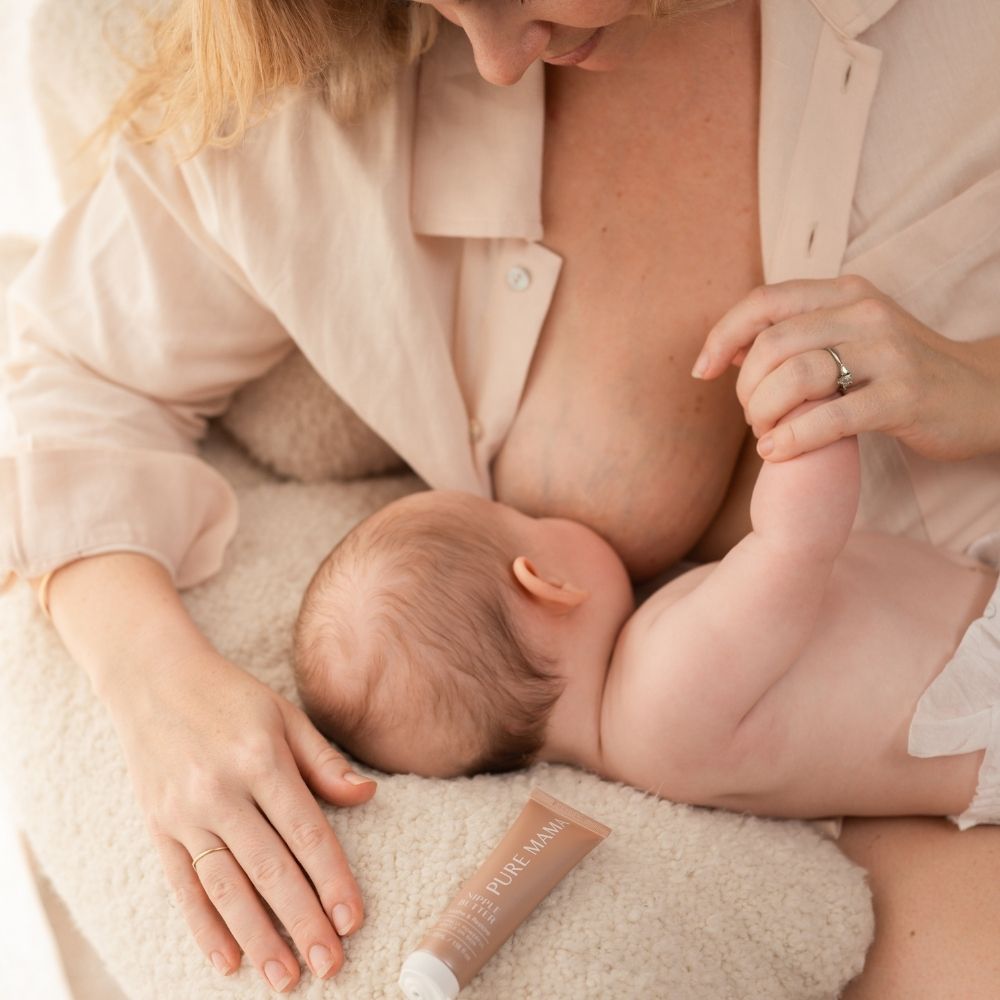 A woman breastfeeds a baby, gently holding its hand. A tube of Nipple Butter by Pure Mama NZ rests beside them on a textured blanket, promising skin nourishment. Both wear soft, neutral tones in a serene atmosphere enhanced by organic butter ingredients.