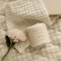 A soft pink flower, a rolled Cotton Muslin Set by Pure Mama NZ, and a white textured blanket sit on a beige plaid quilt in a gently lit, neutral space.