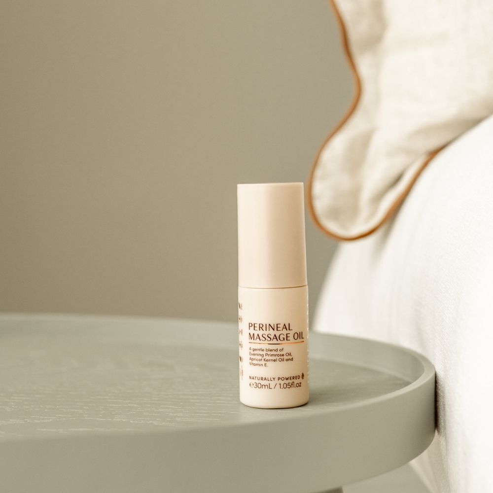 A small bottle of Pure Mama NZ Perineal Massage Oil for birth preparation sits on a round, light gray bedside table next to a bed with light bedding and a pillow with brown piping.