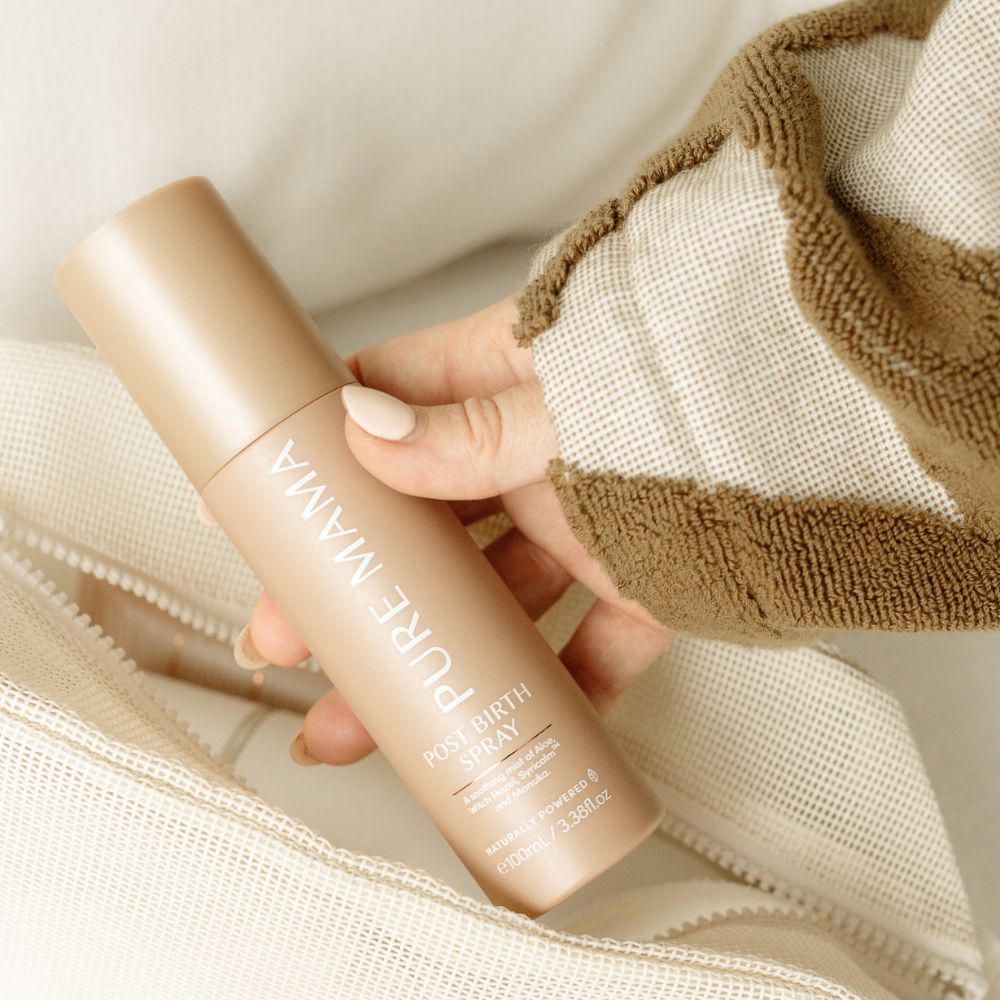 A hand with manicured nails holds a beige bottle from the Pure Mama NZ Birth & Postpartum Recovery Kit above a cream zippered fabric surface. The person is wearing a textured brown and cream long-sleeve top.