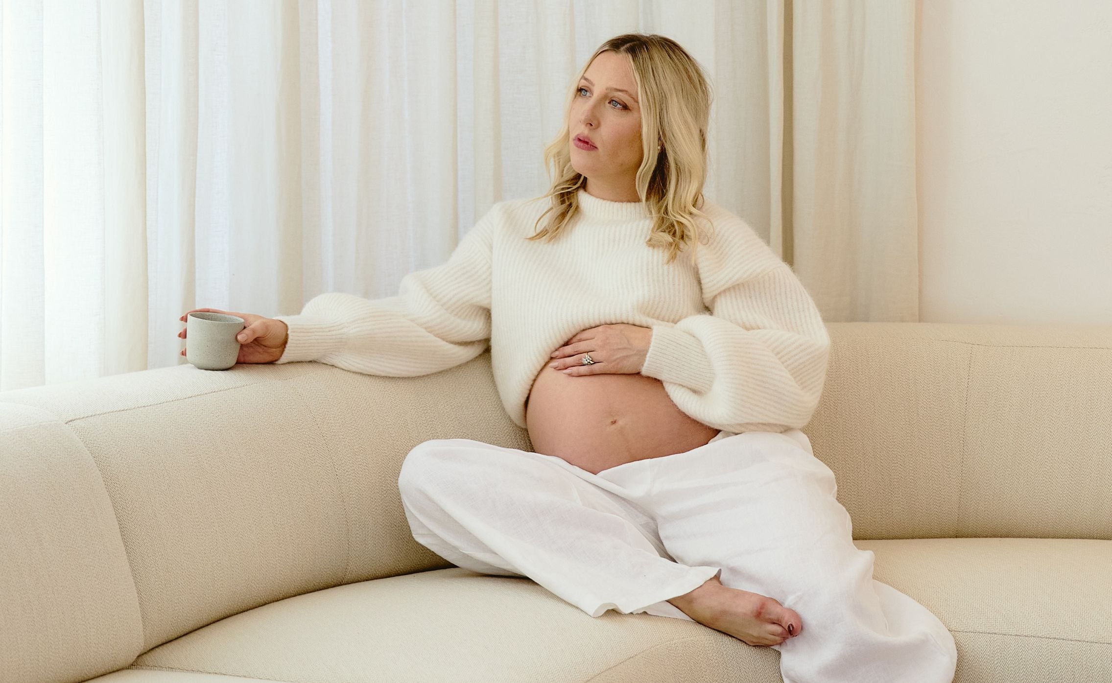 A pregnant woman in a cream sweater and white pants sits on a beige sofa, holding her belly with one hand and a mug in the other, looking thoughtfully out of frame.
