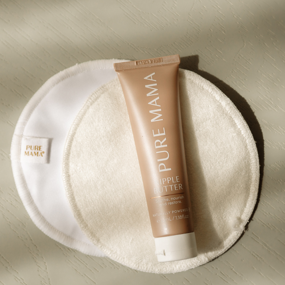 A tube of PURE MAMA Skincare Nipple Butter sits on two PURE MAMA Skincare Nursing Pads (set), arranged on a light, textured surface with sunlight casting gentle shadows.
