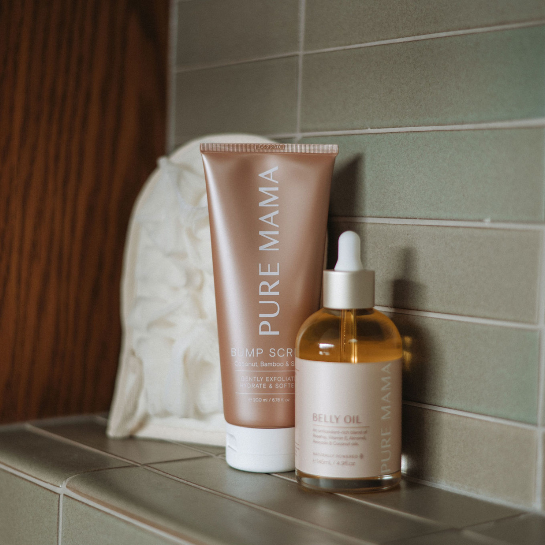 The Bump Love Set by Pure Mama NZ, featuring a Belly Oil bottle with a dropper and a Bump Scrub tube, is elegantly displayed on beige tiles beside a fluffy white bath sponge.