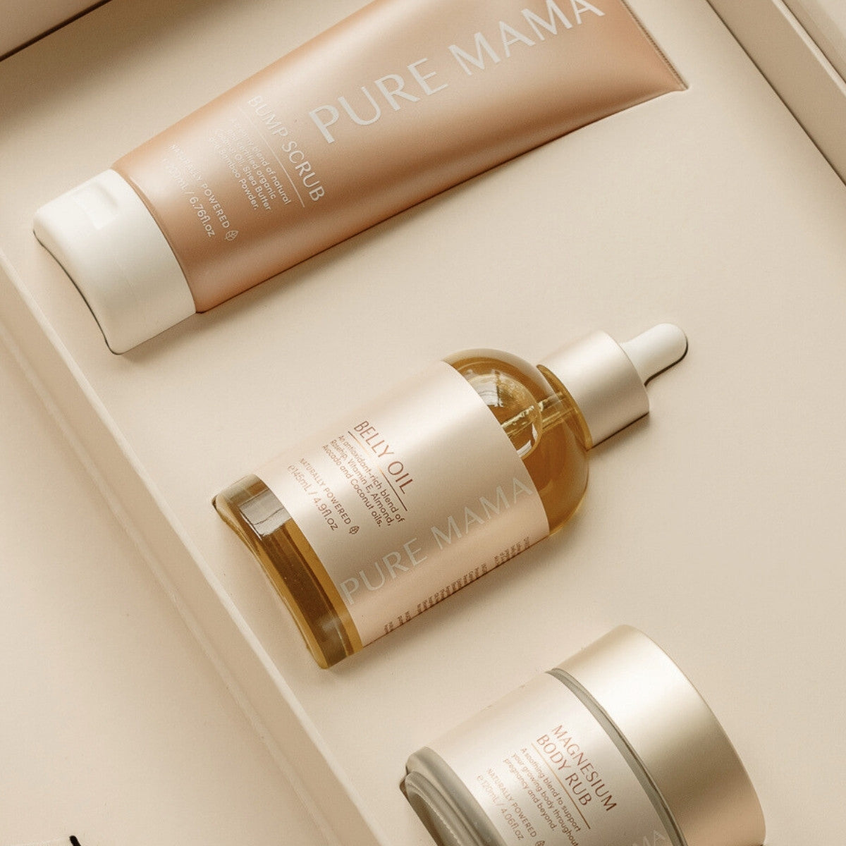 A flat lay on a beige surface showcases Pure Mama NZ essentials: a tube of scrub, the Stretch Mark Belly Oil bottle with a dropper cap, and a jar of body rub, each elegantly labeled in white font.