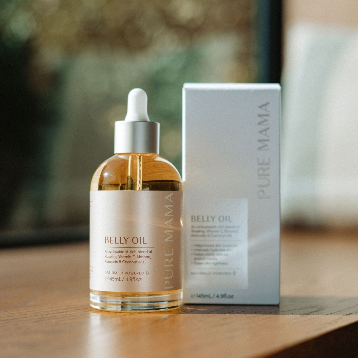 A bottle of Pure Mama NZ Stretch Mark Belly Oil with a dropper rests beside its box on a wooden surface. The label features a blend of avocado and coconut oils, known for enhancing skin elasticity, as sunlight softly illuminates the scene.
