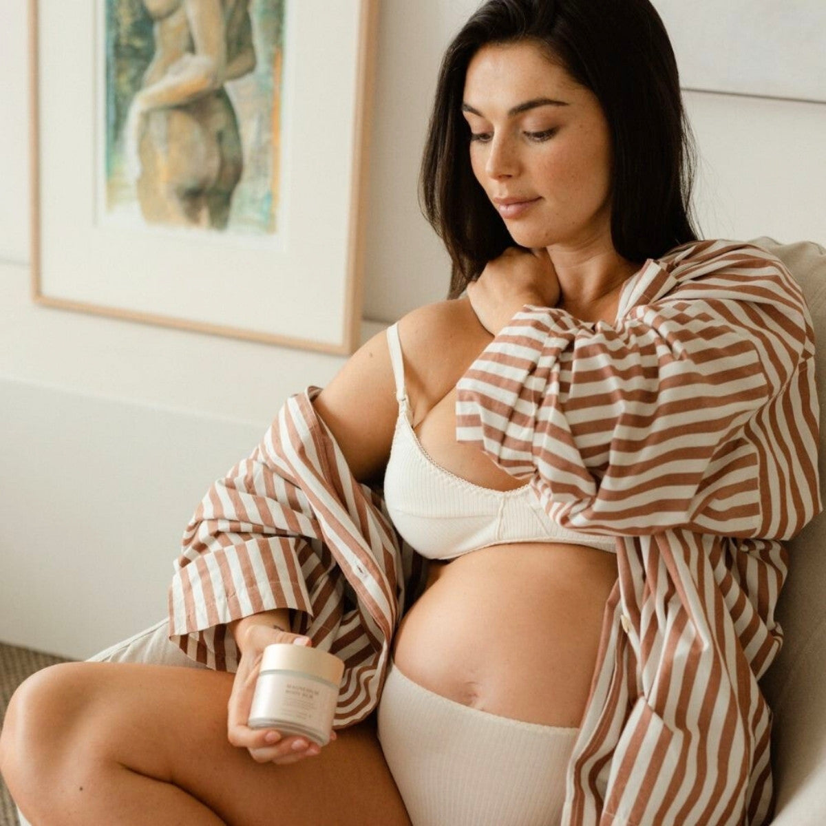 A pregnant woman sits on a chair in a striped robe and white bra, gently holding her stomach. She focuses on stretch mark prevention with Pure Mama NZs The Essentials Set, holding the jar of cream. The rooms muted tones and artwork create an atmosphere of calm and pregnancy support.