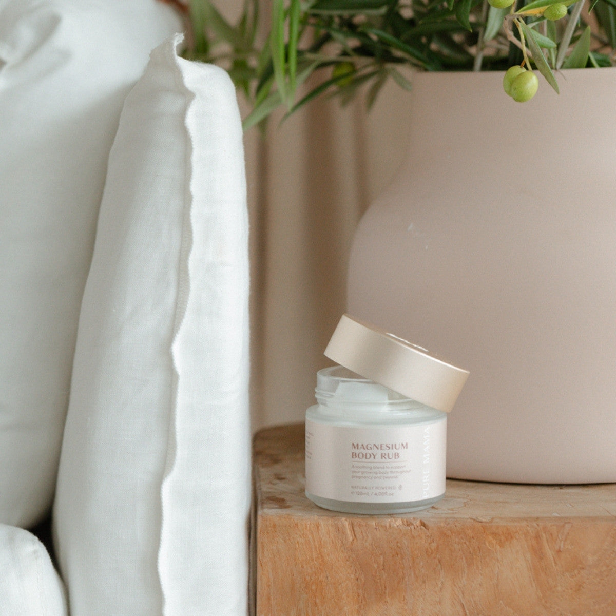 A jar from The Essentials Set by Pure Mama NZ with a beige lid sits on a wooden surface, next to a large tan vase with greenery and a white pillow, creating a serene postpartum self-care ambiance.