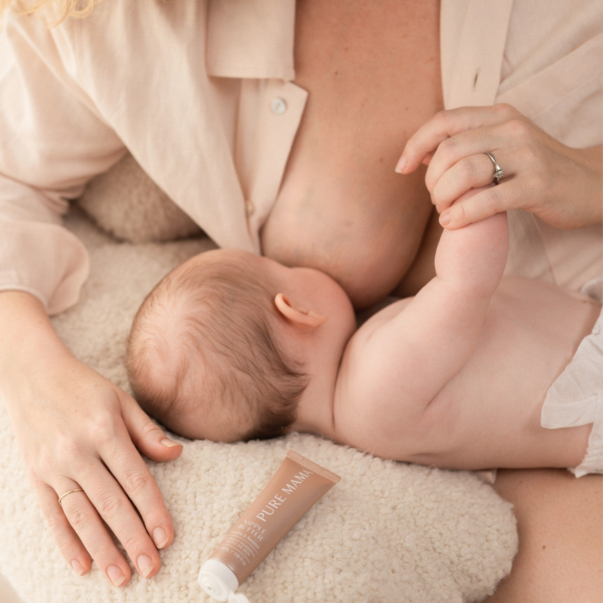 A baby is breastfeeding on a soft blanket while the mother supports with one hand, embracing her postpartum care. Nearby, a tube labeled The Essentials Set from Pure Mama NZ can be seen. Both wear soft, neutral-colored clothing.