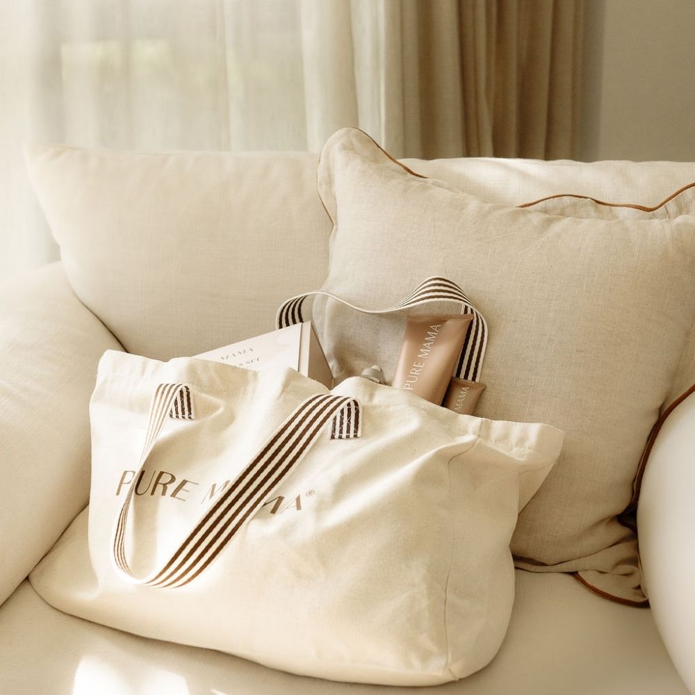A beige Luxe Large Tote Bag by PURE MAMA Skincare sits elegantly on a light sofa with cushions. Available for a limited time, it reveals skincare products inside. Soft natural light filters through a sheer curtain in the background.