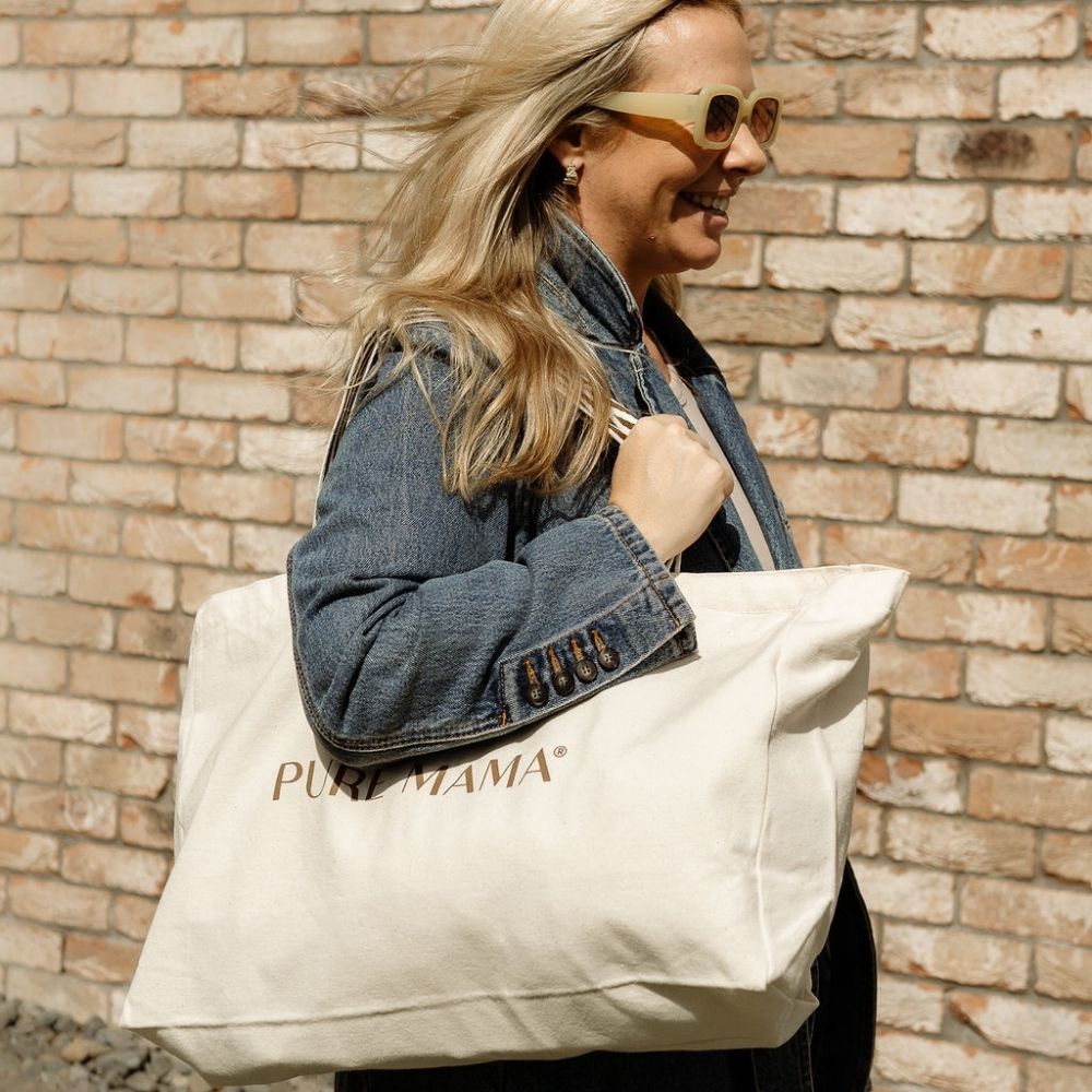 A person in sunglasses and a denim jacket strolls past a brick wall, carrying a Luxe Large Tote Bag branded with PURE MAMA Skincare.