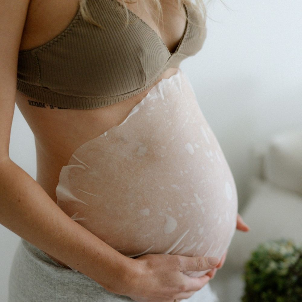 A pregnant person wearing a beige bra and gray pants gently applies the Pure Mama NZ Belly Hydration Mask to their bare belly, caring for stretch marks with both hands. The softly blurred background highlights the nurturing moment.