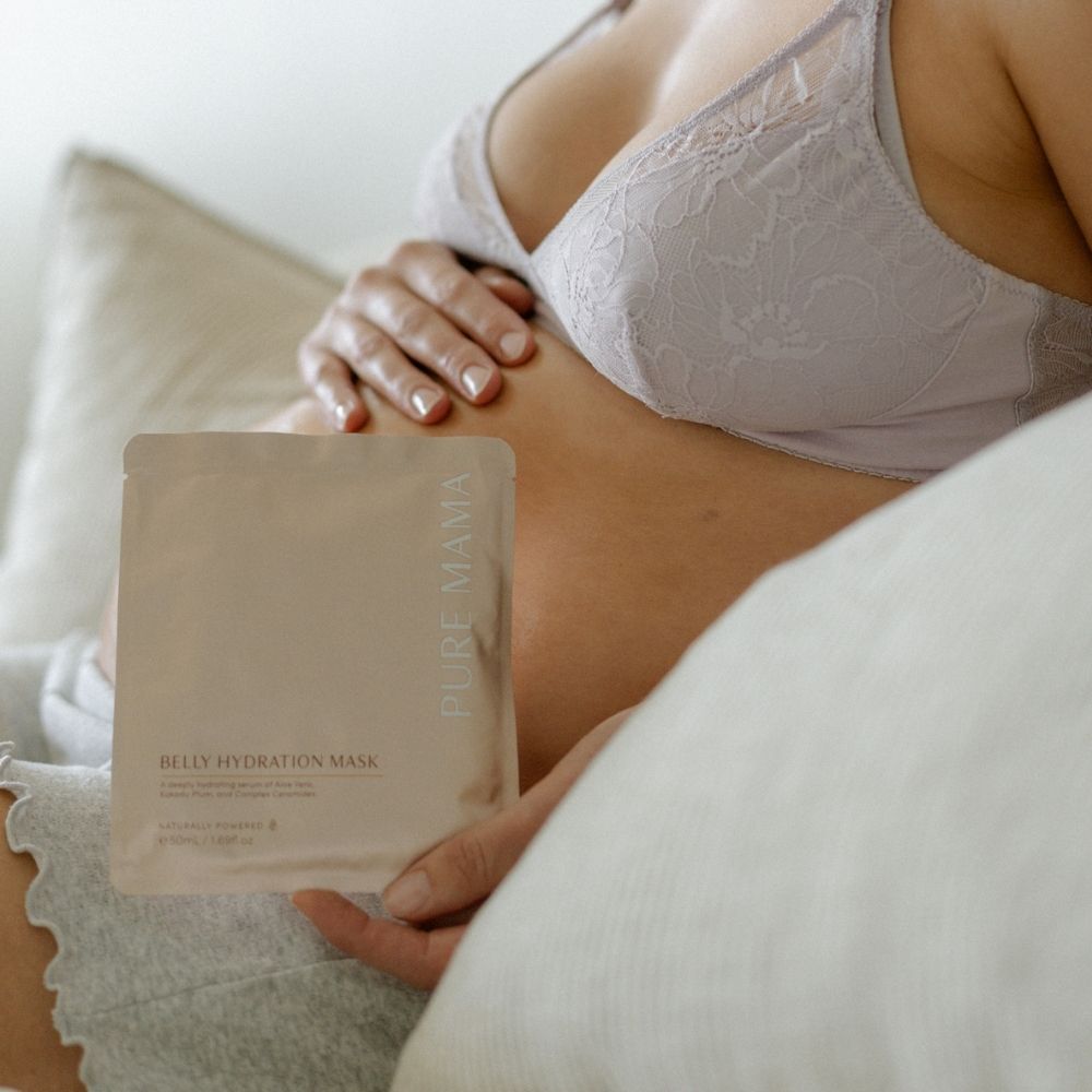 A pregnant person in a light-colored bra relaxes on the couch, holding their belly and a Pure Mama NZ Belly Hydration Mask—enjoying a peaceful moment while supporting their skin barrier with this hydrating sheet mask.