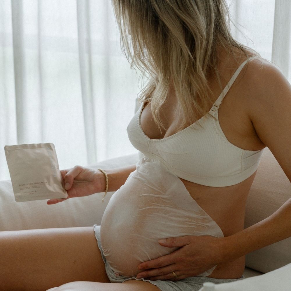 A pregnant woman on a sofa applies the Pure Mama NZ Belly Hydration Mask to her bare belly, holding the product packet as she cares for stretch marks in soft, filtered light.
