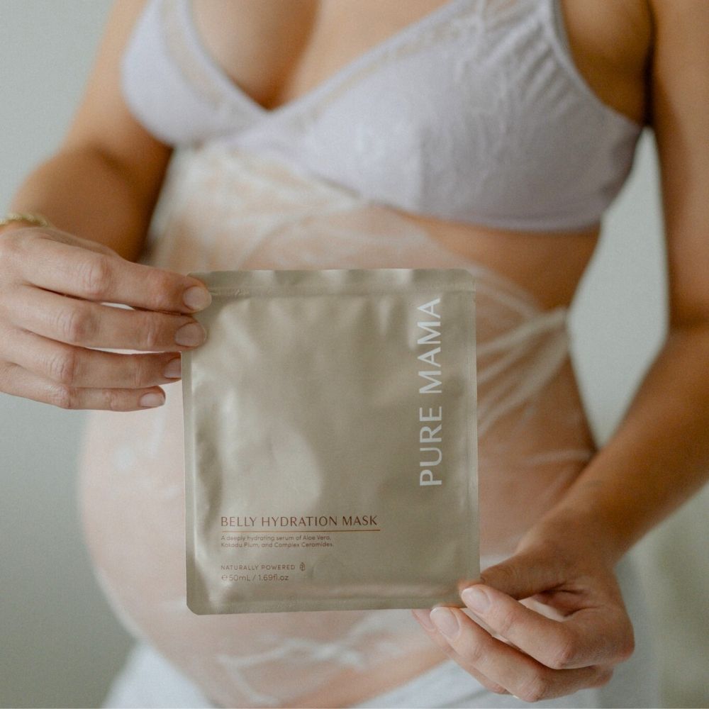 A pregnant person in a light bra holds a gold packet labeled Pure Mama NZ Belly Hydration Mask in front of their belly with white cream marks—ideal for nourishing skin and supporting the skin barrier against stretch marks.