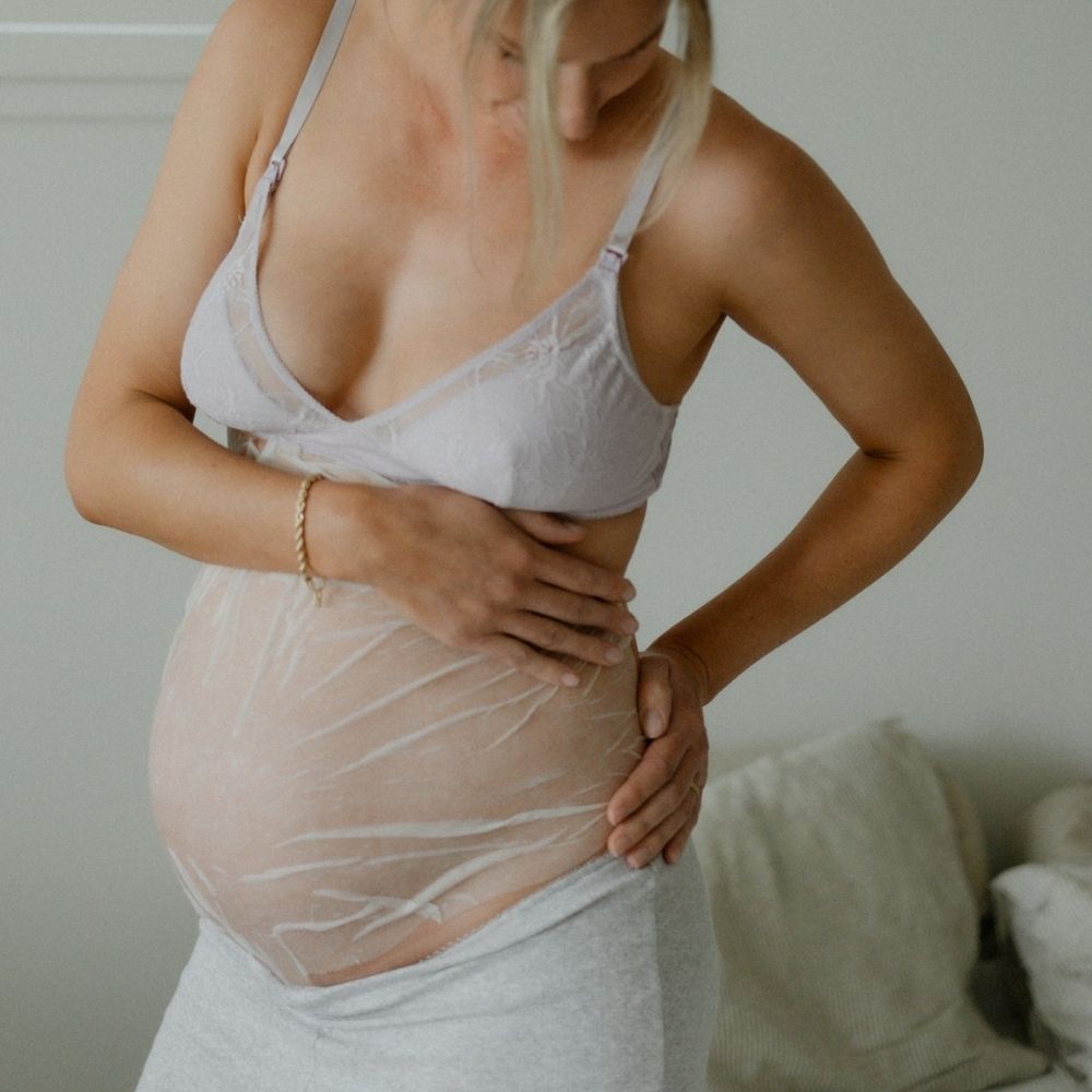 A pregnant woman stands indoors in soft light, gently touching her bare belly covered with Pure Mama NZs Belly Hydration Mask to hydrate her skin and help prevent stretch marks. Shes wearing a bra and gray skirt.