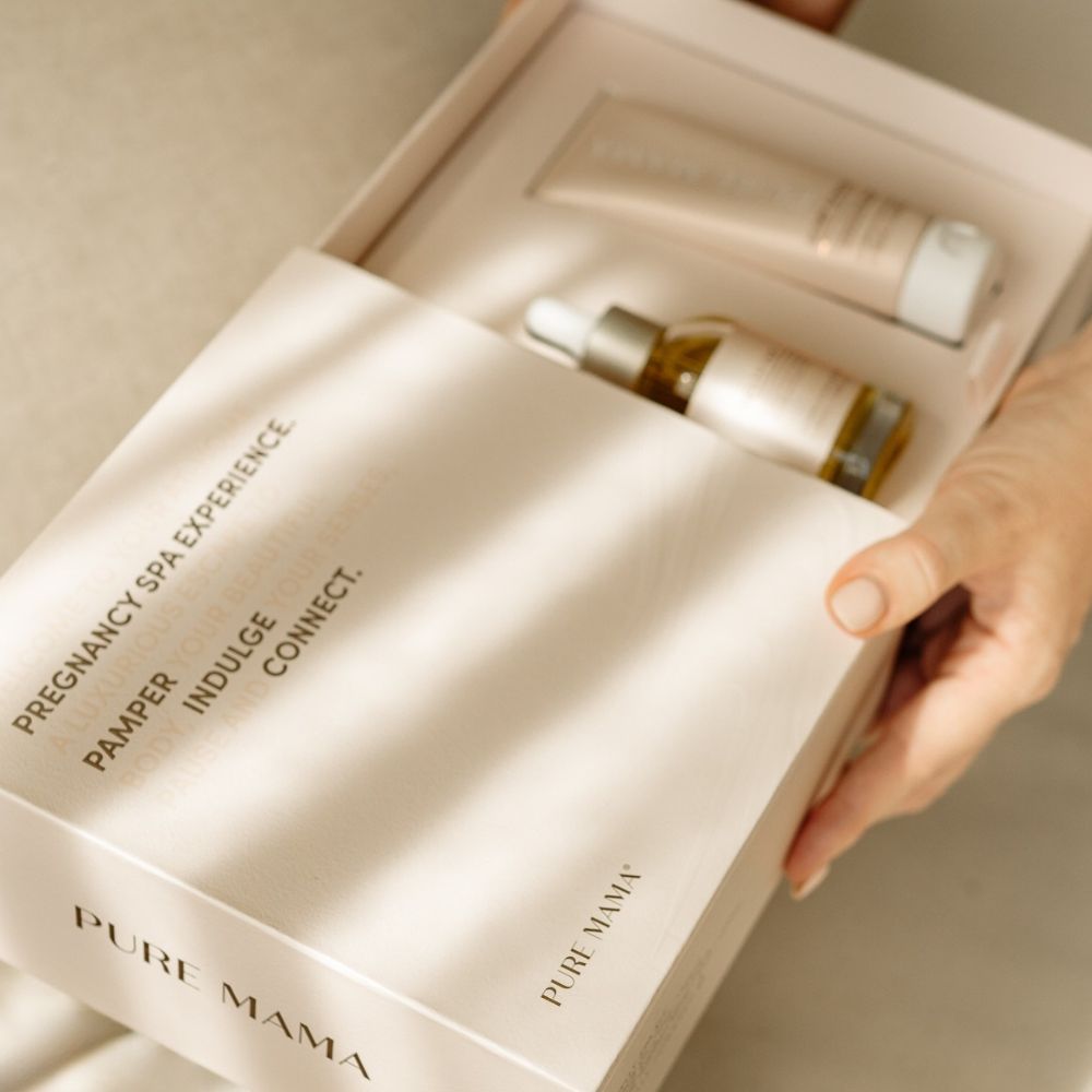 A person holds an open Pure Mama NZ Belly Spa Kit gift box labeled Pregnancy Spa Experience: Pamper. Indulge. Connect. The self-care set features skincare products, including a tube and a glass bottle with a dropper.