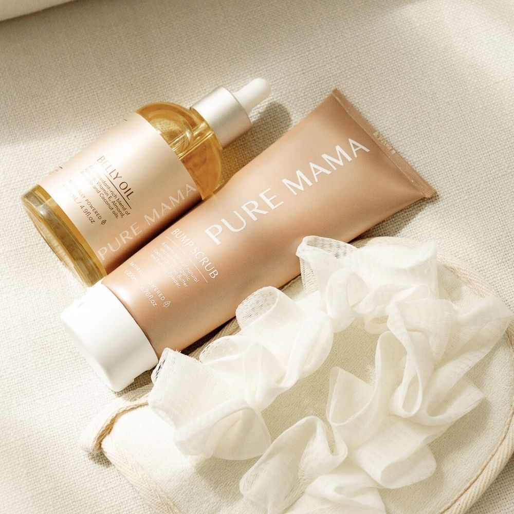 The Pure Mama NZ Bump Love Set, featuring Belly Oil and Belly Rub, is displayed on soft beige fabric beside a white shower pouf—ideal for stretch mark prevention in your skincare routine.