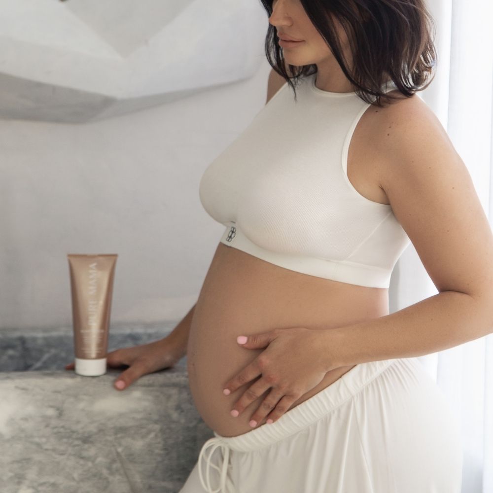 A pregnant woman in a white crop top and pants stands by a counter, gently touching her belly. The Pure Mama NZ Bump Love Set is placed on the counter next to her against a softly lit, neutral background.