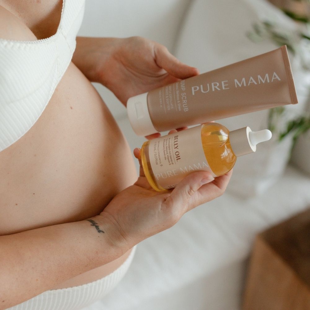 A person in a white top holds Pure Mama NZs Bump Love Set, featuring a tube of Bump Scrub and a bottle of Belly Oil, promising stretch mark prevention. The pregnant individual stands with plants and a wooden table softly blurred in the background.