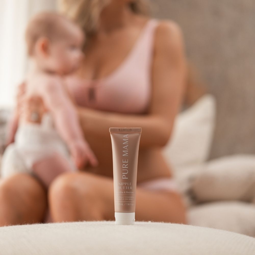 In a cozy, softly-lit setting, a Pure Mama NZ nipple butter from the Mama & Baby Set is in focus. In the background, a woman in pink cradles a baby.