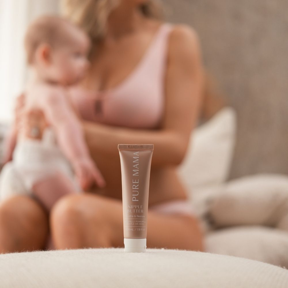 A tube of Nipple Butter by Pure Mama NZ sits in focus on a soft surface, while a woman in pink, breastfeeding her baby, is blurred in the cozy background. The organic formula provides gentle skin nourishment for new mothers.