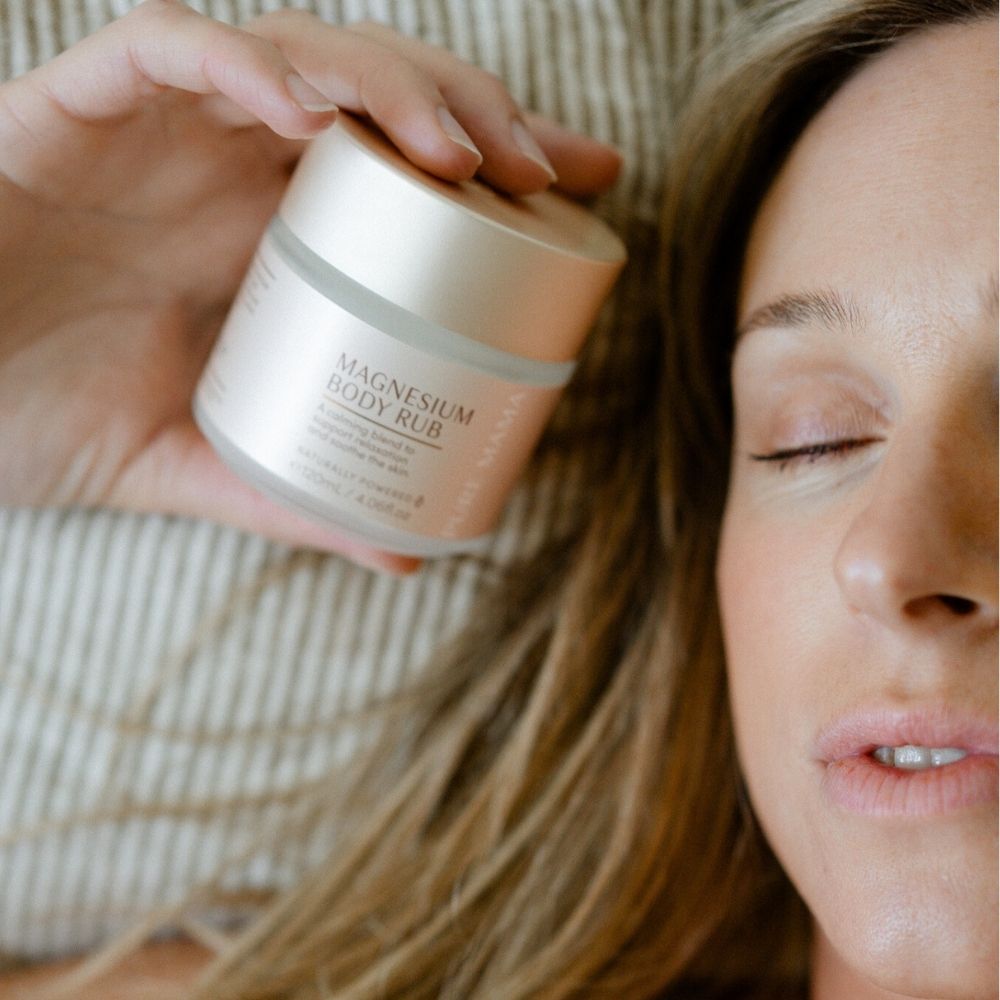 A woman with closed eyes relaxes on a pillow, holding a jar from The Essentials Set by Pure Mama NZ close to her face, highlighting relaxation and skincare as key elements of her self-care routine.