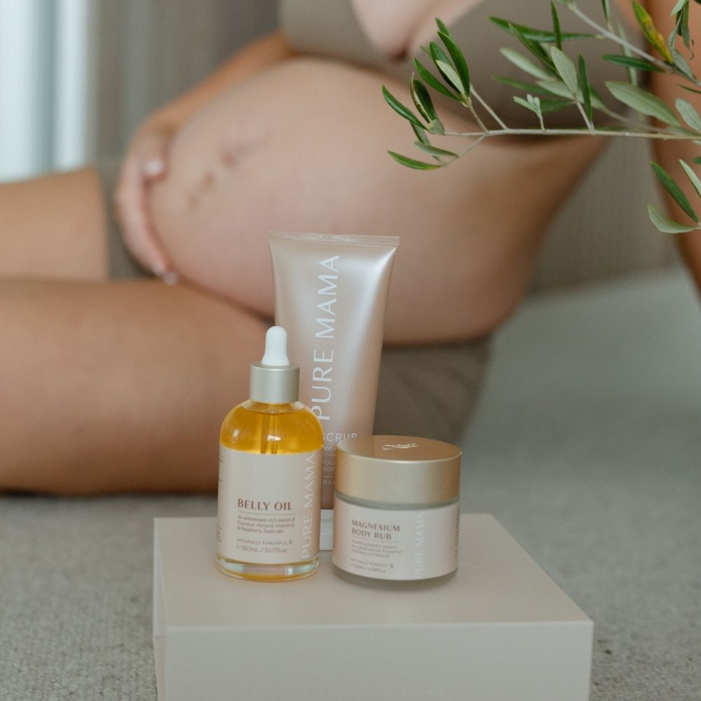 A pregnant person sits in the background, showcasing the beauty of motherhood. In the foreground, a Pure Mama NZ Pregnancy Care Set is displayed on a box: it includes a dropper bottle labeled Belly Oil, a tube labeled Scrub, and a jar of nourishing Magnesium Body Rub.