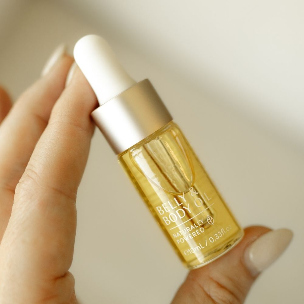A hand holds a Belly Oil Mini by Pure Mama NZ, perfect for enhancing skin elasticity. The small glass bottle, featuring a metallic cap and dropper, contains yellow oil with the label Naturally Powered against a blurred background.
