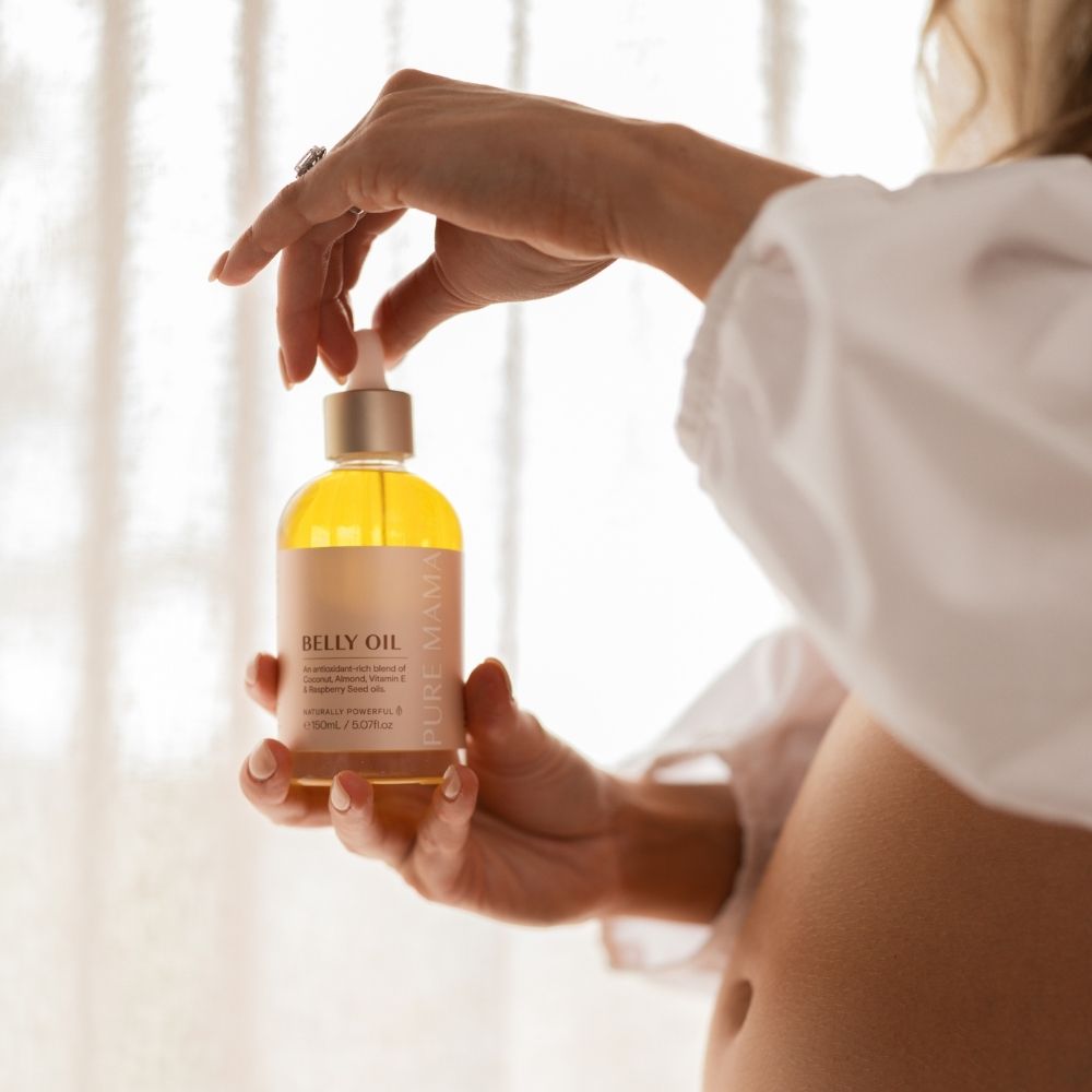 A person holds a bottle of Pure Mama NZs Belly Oil Mini with a dropper in front of a softly lit sheer curtain. The label notes its promise to enhance skin elasticity and reduce stretch marks. Theyre wearing a white shirt, revealing part of their abdomen and capturing the products essence.