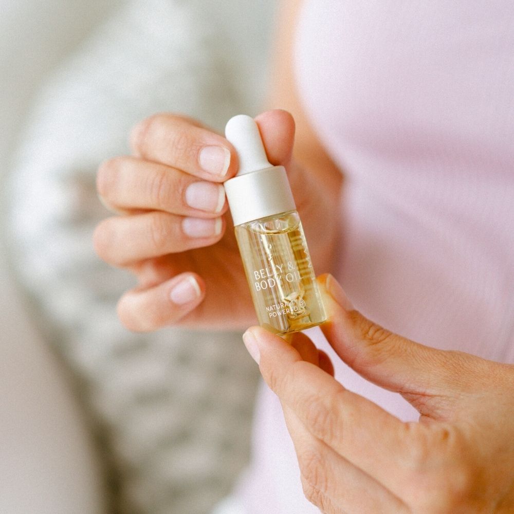 A person in a light pink top holds a Pure Mama NZ Belly Oil Mini dropper bottle, ideal for improving skin elasticity. The bottle contains a light-colored liquid against a softly blurred background.