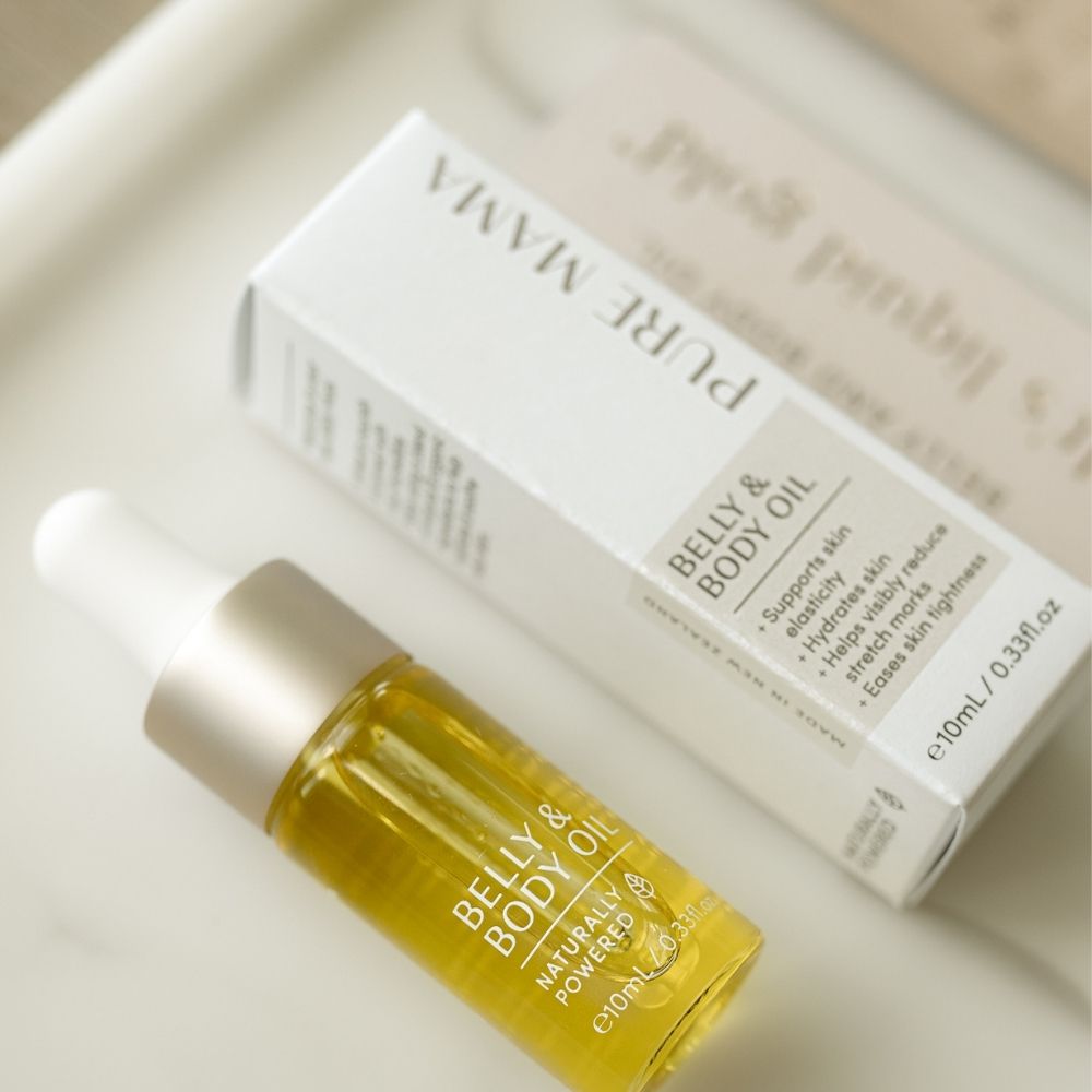 A bottle of Pure Mama NZs Belly Oil Mini with a white dropper cap is on a tray beside its box, which highlights benefits like skin support and elasticity for pregnancy skincare. This natural, golden-yellow formula comes in a 10ml volume.