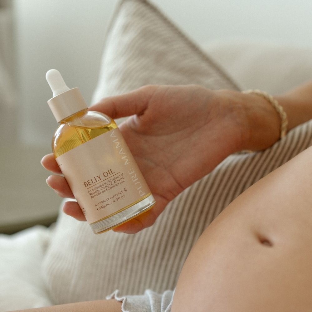 A hand holds a bottle of Pure Mama NZ Stretch Mark Belly Oil with a dropper top, next to a pregnant belly—perfect for pregnancy care and supporting skin elasticity. A beige pillow is visible in the background.