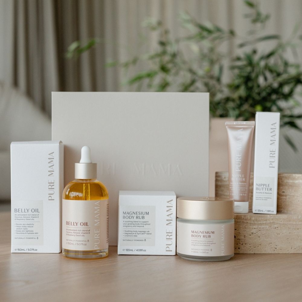 Pure Mama NZs The Essentials Set, featuring belly oil, magnesium body rub, and nipple butter in minimalist white packaging, is displayed on a table with a blurred plant backdrop enhancing its serene vibe.